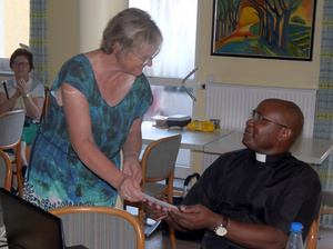 Father Dunstan sitzt an einem Tisch. Links daneben steht Hiltrud Altenhöfer. Sie beugt sich zu ihm freundlich hinunter und übergibt einen Umschlag. 